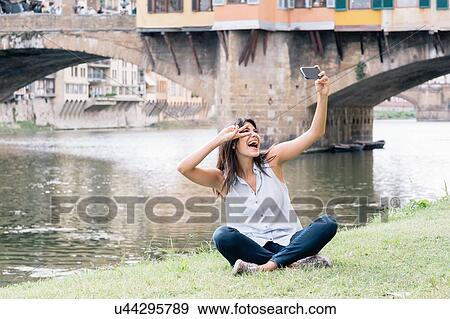 Stock Photo - Young woman sitting on riverbank in front of Ponte Vecchio and Arno river using smartphone to take selfie, Florence, Tuscany, Italy. Fotosearch
