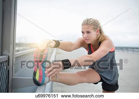 Stock Image - Young woman stretching leg against railing, outdoors. Fotosearch