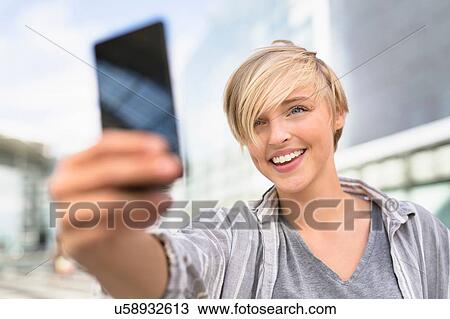 Stock Image - Young woman taking smartphone selfie in city. Fotosearch