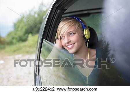 Stock Image - Young woman wearing headphones looking out of open car window. Fotosearch