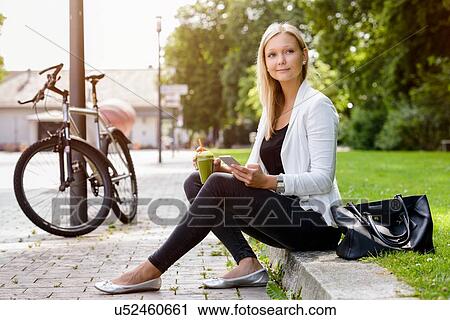 Stock Image - Young woman with smoothie drink and smartphone sitting in park. Fotosearch