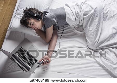 Stock Photo - MODEL RELEASED. Young man asleep in bed with laptop and phone.. Fotosearch