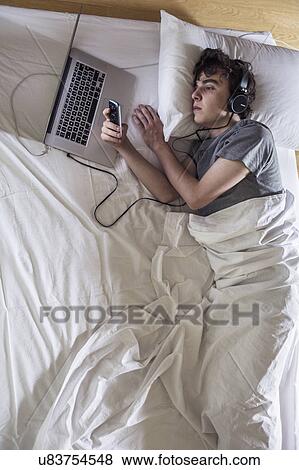 Stock Photo - MODEL RELEASED. Young man in bed with laptop and phone.. Fotosearch