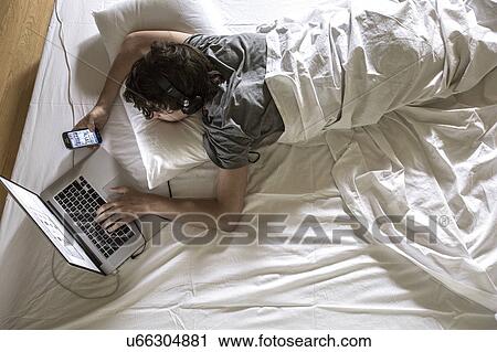 Stock Image - MODEL RELEASED. Young man lying in bed using laptop and phone.. Fotosearch