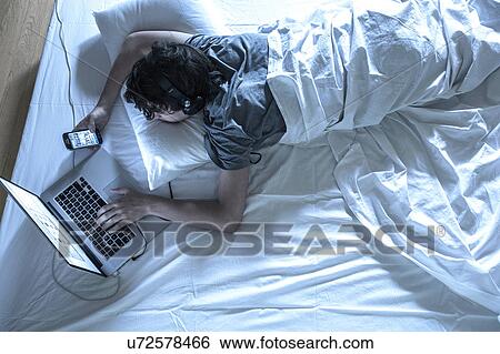 Stock Photograph - MODEL RELEASED. Young man lying in bed using laptop and phone.. Fotosearch