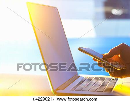Stock Photo - Business man at laptop computer with smartphone in office with sunlight coming through window. Fotosearch