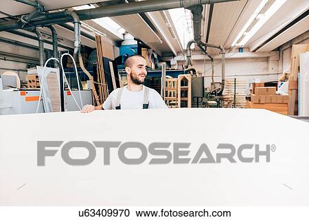 Stock Image - Carpenter with large sheet of wood in workshop. Fotosearch