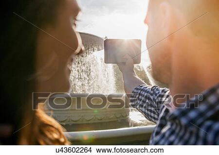 Picture - Couple taking selfie on phone, fountain in background. Fotosearch