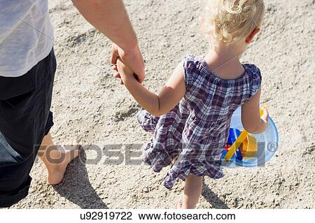 Stock Image - Father and female toddler strolling on beach with toy bucket. Fotosearch