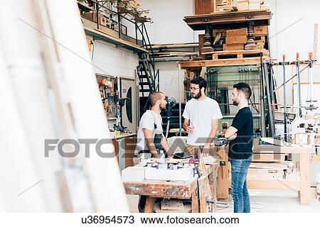 Stock Image - Friends standing around workbench in carpentry workshop talking. Fotosearch