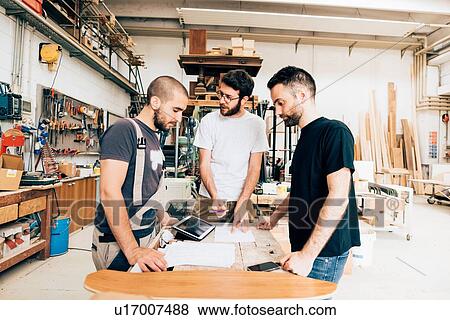 Stock Photo - Friends standing around workbench in carpentry workshop talking. Fotosearch