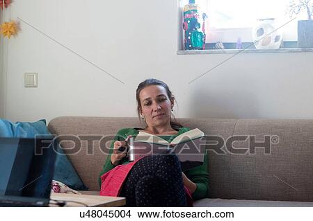 Front view of mid adult woman sitting on sofa looking down reading book View Large Photo Image Picture - Front view of mid adult woman sitting on sofa looking down reading book. Fotosearch