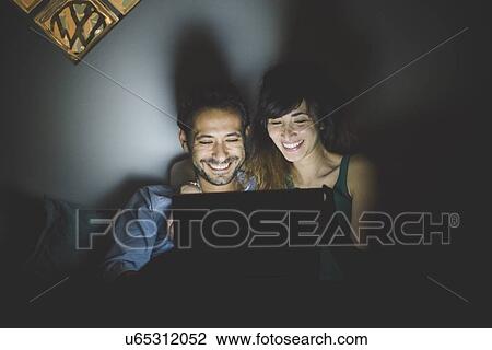 Front view of young couple illuminated by laptop computer screen, looking down smiling View Large Photo Image Stock Image - Front view of young couple illuminated by laptop computer screen, looking down smiling. Fotosearch