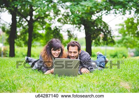 Stock Image - Front view of young couple lying on front on grass using laptop computer smiling. Fotosearch