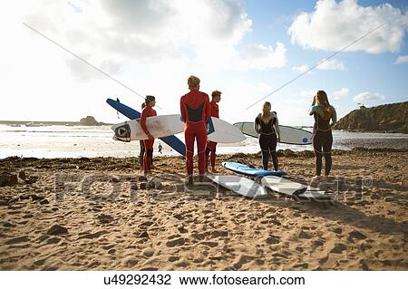 Stock Image - Group of surfers standing on beach, holding surfboards, rear view. Fotosearch