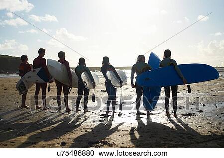Stock Image - Group of surfers, standing on beach, holding surfboards, rear view. Fotosearch
