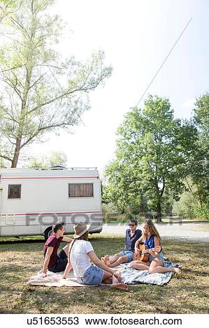 Stock Image - Group of young adults sitting on picnic blanket, relaxing, camper van in background. Fotosearch