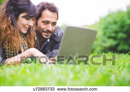 Stock Image - Head and shoulders of young couple lying on front on grass using laptop computer smiling. Fotosearch