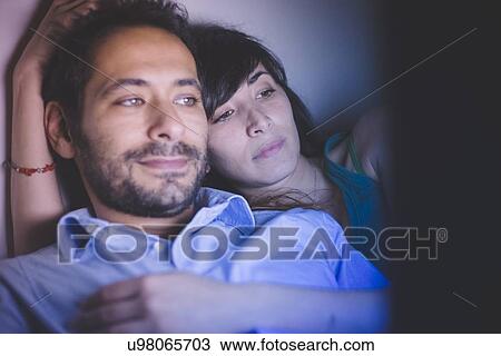 Stock Image - Head and shoulders of young couple lying down illuminated by laptop computer screen. Fotosearch