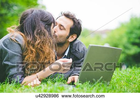 Stock Photograph - Head and shoulders of young couple lying on front on grass with laptop computer smiling. Fotosearch