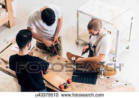 Stock Image - High angle view of young men standing around workbench attaching wheels to skateboards. Fotosearch
