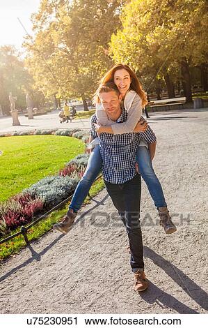 Stock Image - Man giving woman piggy back in park. Fotosearch