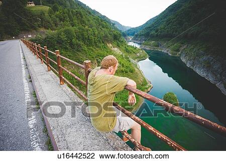 Stock Image - Man sitting on railings by road, Lake Garda, Italy. Fotosearch