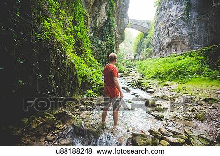 Stock Photo - Man standing in stream, rock hills and stone bridge in background, Garda, Italy. Fotosearch