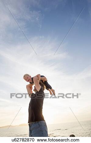 Stock Image - Mature man lifting up his toddler daughter on beach, Calvi, Corsica, France. Fotosearch