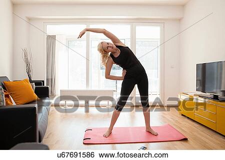 Stock Photograph - Mature woman in lounge standing on yoga mat bending over sideways arm raised stretching. Fotosearch