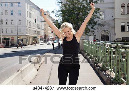 Mature woman outdoors wearing sports clothing arms raised looking down smiling View Large Photo Image Stock Photo - Mature woman outdoors wearing sports clothing arms raised looking down smiling. Fotosearch