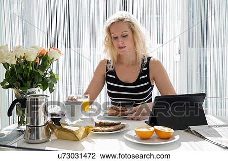 Mature woman sitting at table with digital tablet looking down eating breakfast View Large Photo Image Stock Image - Mature woman sitting at table with digital tablet looking down eating breakfast. Fotosearch