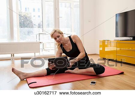 Stock Photo - Mature woman sitting on yoga mat wearing earbuds holding digital tablet. Fotosearch