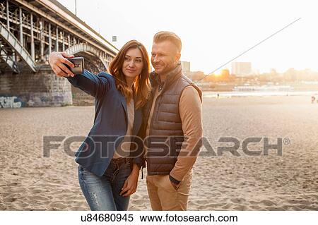 Stock Photography - Mid adult couple by bridge taking selfie on phone. Fotosearch