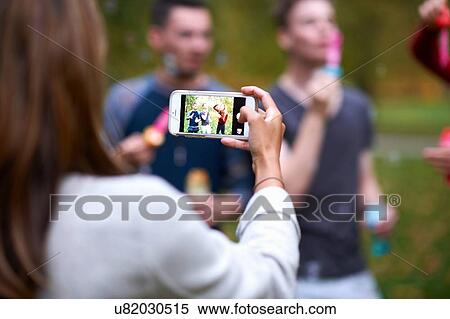 Over shoulder view of young woman taking smartphone photograph of friends in park View Large Photo Image Stock Photography - Over shoulder view of young woman taking smartphone photograph of friends in park. Fotosearch
