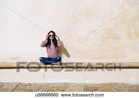 Stock Image - Portrait of young woman adjusting sunglasses on bench, Florence, Italy. Fotosearch
