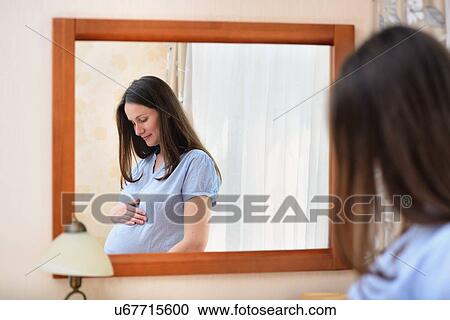 Pregnant woman holding stomach, standing in front of mirror View Large Photo Image Stock Image - Pregnant woman holding stomach, standing in front of mirror. Fotosearch