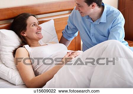 Stock Image - Pregnant woman lying in bed, husband sitting beside her. Fotosearch