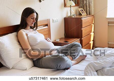 Pregnant woman sitting on bed, holding stomach View Large Photo Image Stock Image - Pregnant woman sitting on bed, holding stomach. Fotosearch