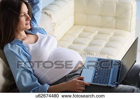 Stock Image - Pregnant woman sitting on sofa, using laptop. Fotosearch