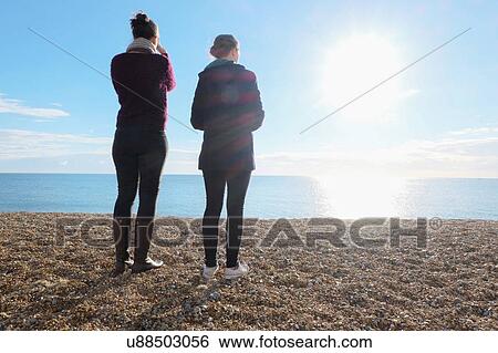 Stock Photograph - Rear view of two young adult sisters looking out at sunlit sea. Fotosearch