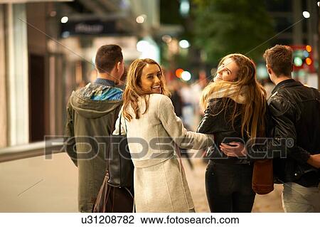 Stock Image - Rear view of two young couples strolling arm in arm along street at night, London, UK. Fotosearch