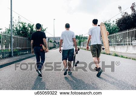 Stock Image - Rear view of young men walking on path carrying skateboards. Fotosearch