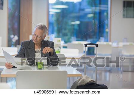 Stock Image - Senior businesswoman with wheeled suitcase reading menu in hotel dining room. Fotosearch