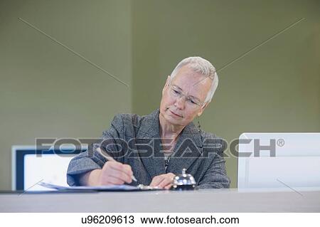 Stock Image - Senior female receptionist writing at hotel reception desk. Fotosearch