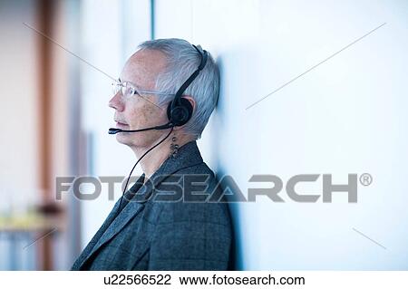 Stock Image - Side view of mature woman leaning against wall wearing telephone headset looking away. Fotosearch