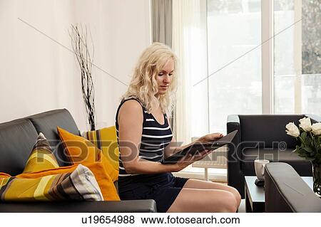 Stock Photo - Side view of mature woman sitting on sofa holding digital tablet looking down. Fotosearch