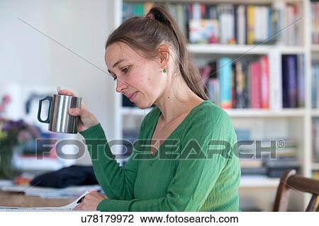 Stock Image - Side view of mid adult women sitting at table reading magazine holding cup. Fotosearch