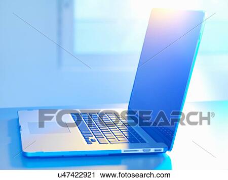 Stock Image - Side view of open laptop on desk, sunlight coming through window. Fotosearch