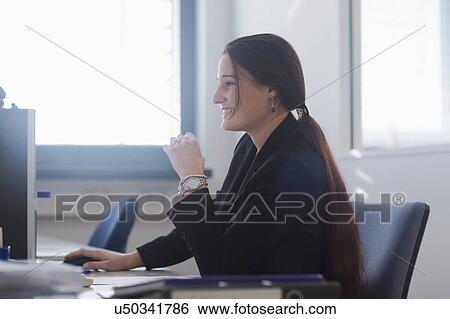 Stock Photograph - Side view of young woman in office sitting at desk using computer smiling. Fotosearch